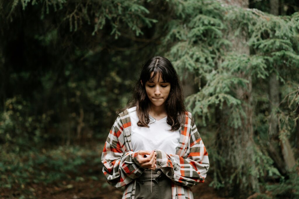 A young girl stands thoughtfully in a serene forest, surrounded by lush greenery.