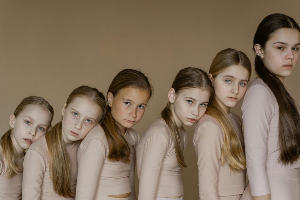 Young ballet students pose together in beige outfits against a brown background.